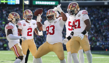 Four San Francisco 49ers players celebrate on the football field, with one player holding the ball and raising his hands in excitement.