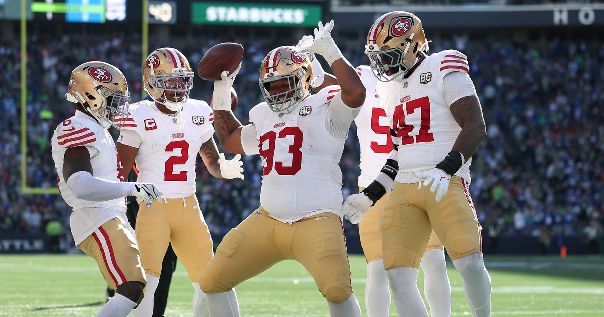 Four San Francisco 49ers players celebrate on the football field, with one player holding the ball and raising his hands in excitement.