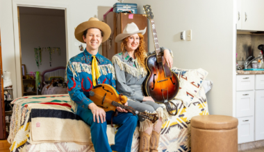 A man and woman dressed in vintage western attire sit on a bed, holding a mandolin and guitar, with a colorful patterned blanket beneath them.