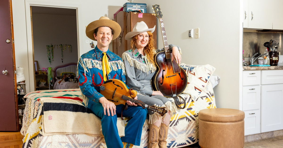 A man and woman dressed in vintage western attire sit on a bed, holding a mandolin and guitar, with a colorful patterned blanket beneath them.