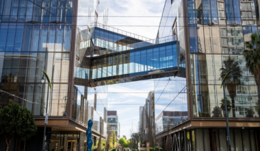 Two modern glass office buildings face each other, connected by a glass skybridge, with trees, sculptures, and a street lined with crosswalks.