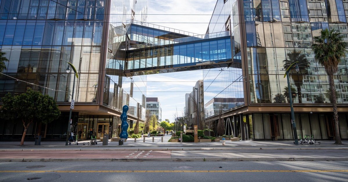 Two modern glass office buildings face each other, connected by a glass skybridge, with trees, sculptures, and a street lined with crosswalks.