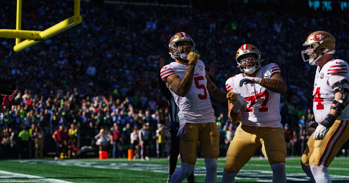Three San Francisco 49ers football players celebrate in the end zone under the goalpost during a game with a crowd in the background.