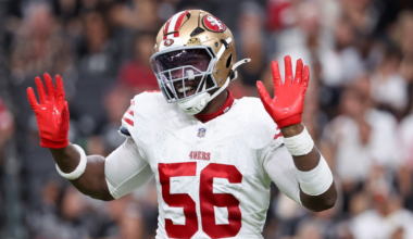 A football player wearing a San Francisco 49ers uniform with the number 56 raises both hands, showing bright red gloves.