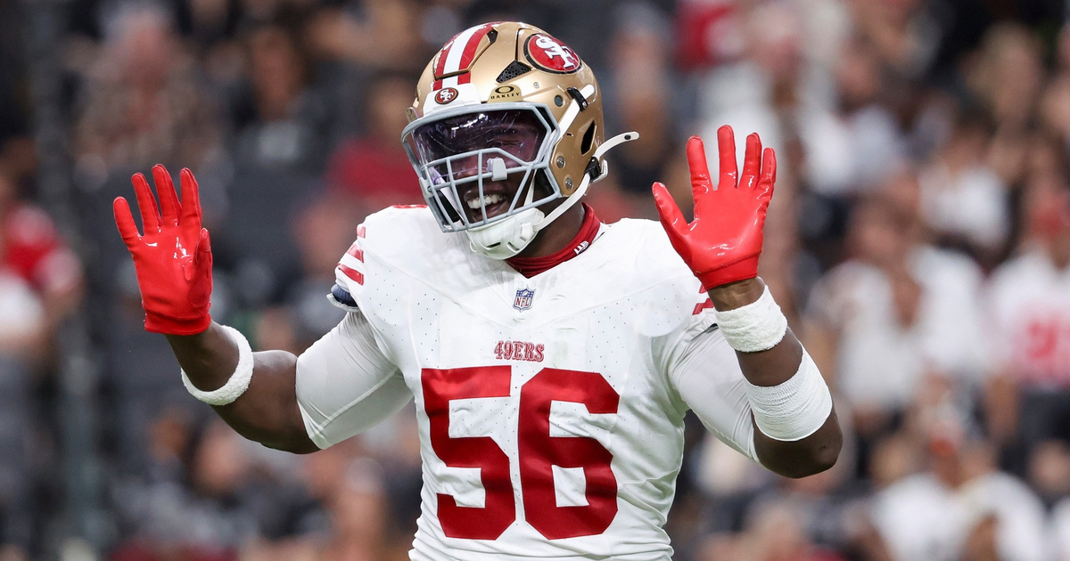 A football player wearing a San Francisco 49ers uniform with the number 56 raises both hands, showing bright red gloves.