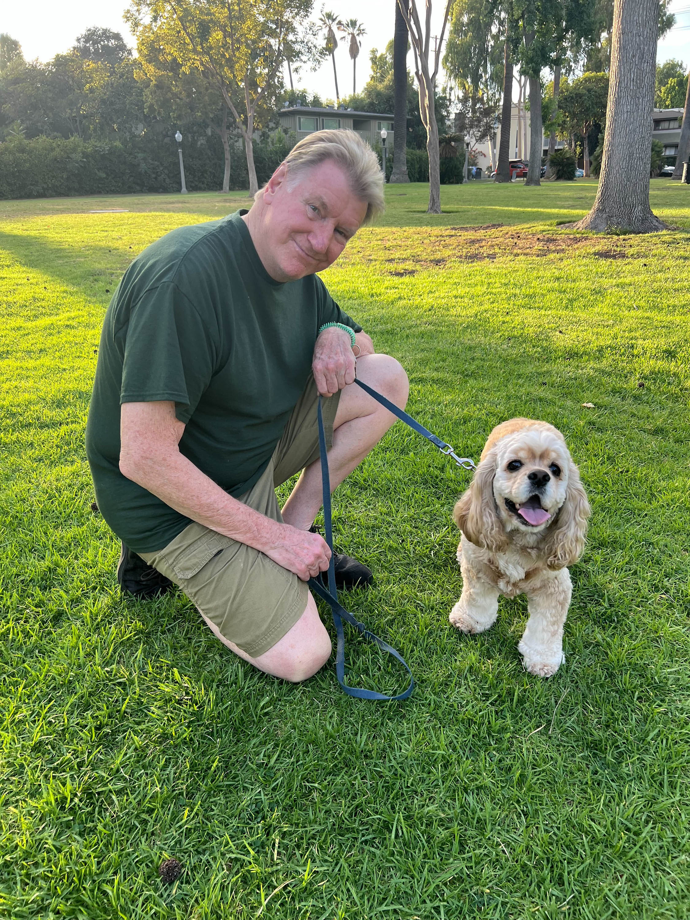 A middle-aged man kneels on lush green grass with his medium-sized dog on a leash beside him.