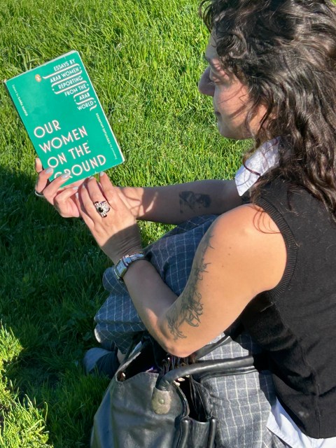 A person sitting on grass reading the book “Our Women on the Ground: Essays by Arab Women Reporting from the Arab World.”
