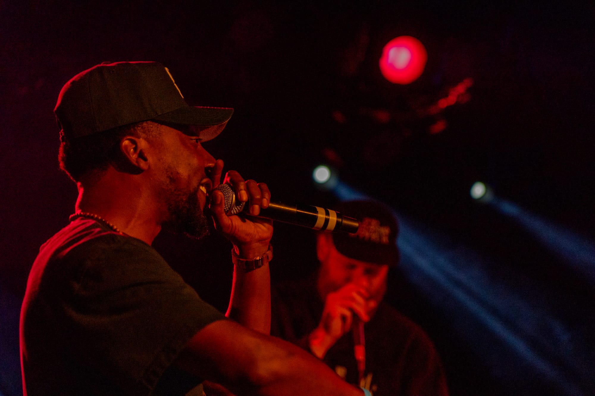 A man in a black hat and shirt holds a microphone in red lighting.