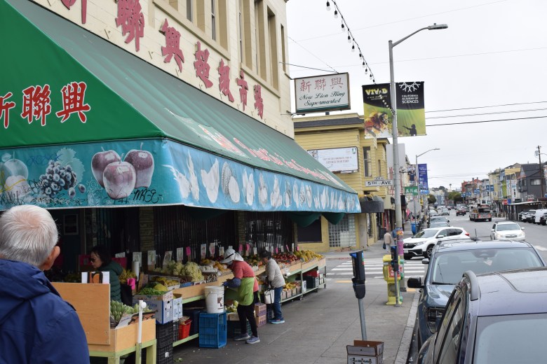 Street view of a market with green awning displaying fruit and Chinese characters; people select produce from outdoor displays, and cars are parked along the sidewalk.
