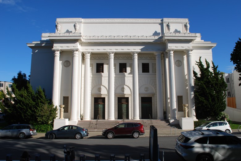 Large white neoclassical building with tall columns and three entrance doors, flanked by trees; several parked cars in front on a sunny day.