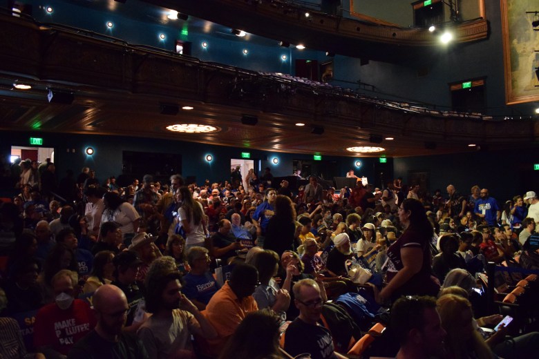 A large crowd of people fills a theater auditorium, seated and standing, as they wait for an event to begin.