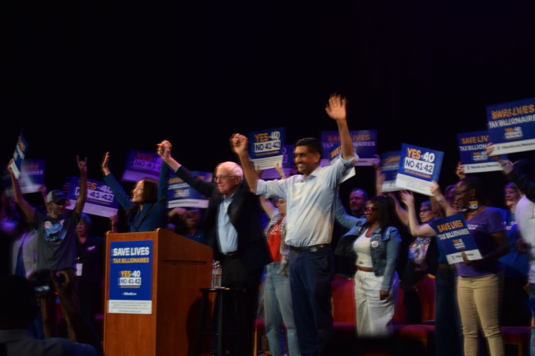 A group of people stand on a stage holding signs that support ballot measures, while some raise their arms and cheer beside a podium.