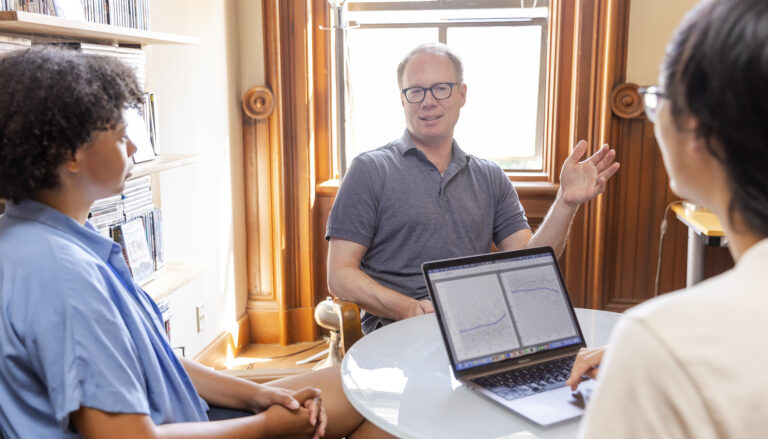 UC Berkeley Associate Professor David Bamman speaks with students seated around a table with a laptop displaying data graphs.