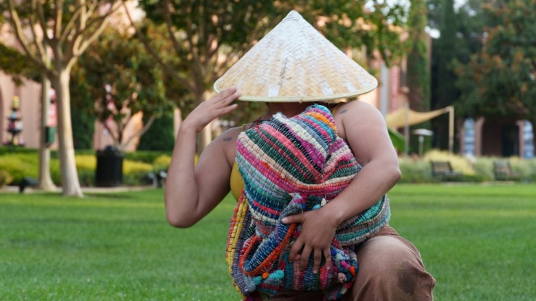 Person in a conical woven hat holding a brightly colored striped textile on a grassy lawn.