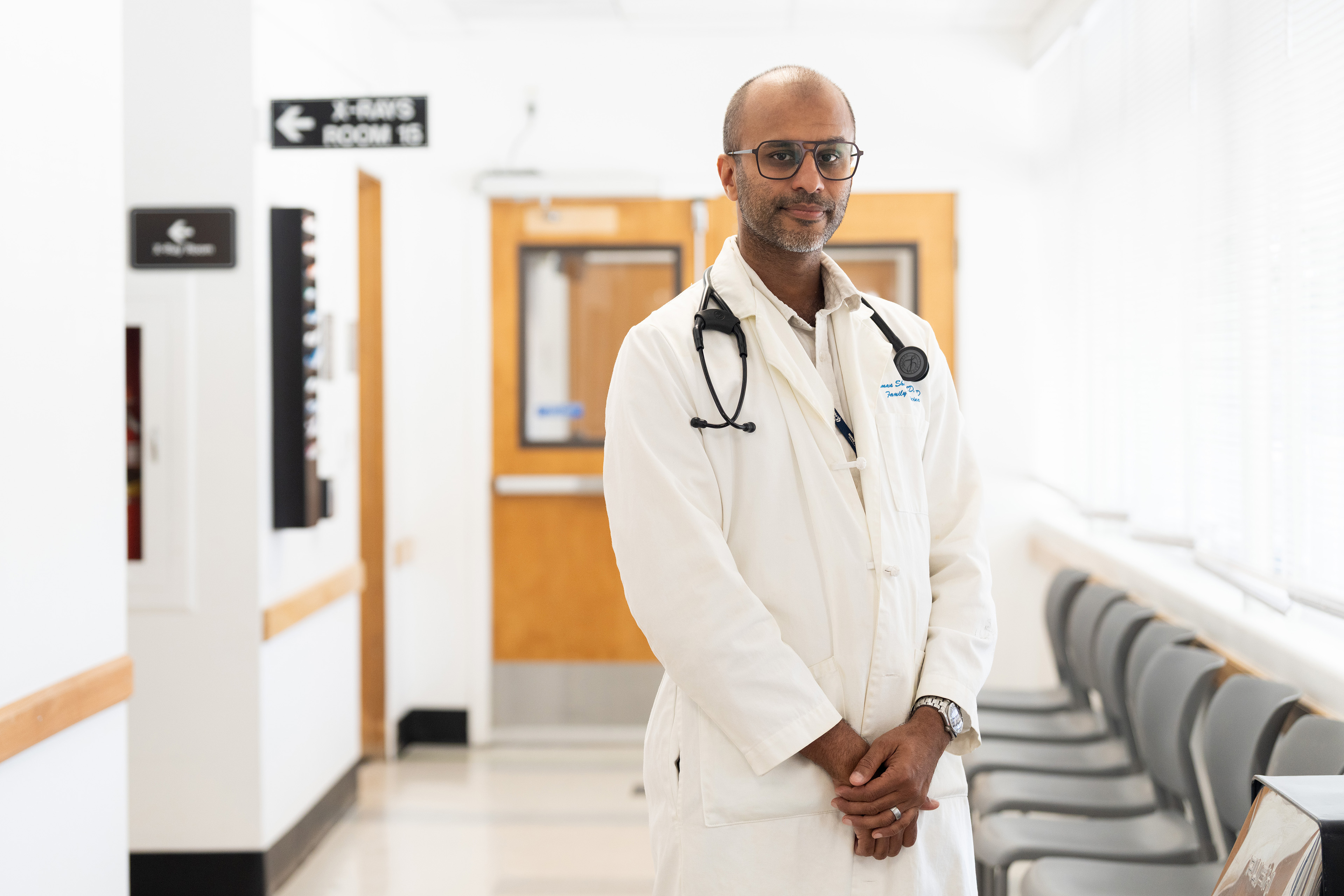 A doctor wearing a white coat and glasses, with a stethoscope around his neck, stands in a medical clinic hallway