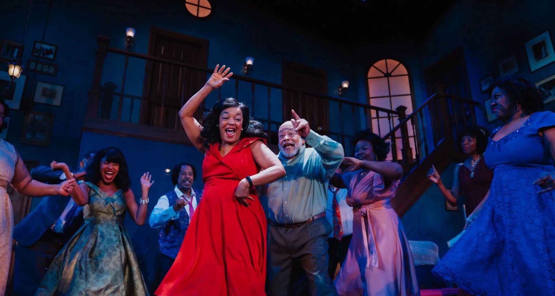 A Black woman in a bright red dress smiles and dances in a dimly lit room with several others