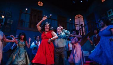 A Black woman in a bright red dress smiles and dances in a dimly lit room with several others