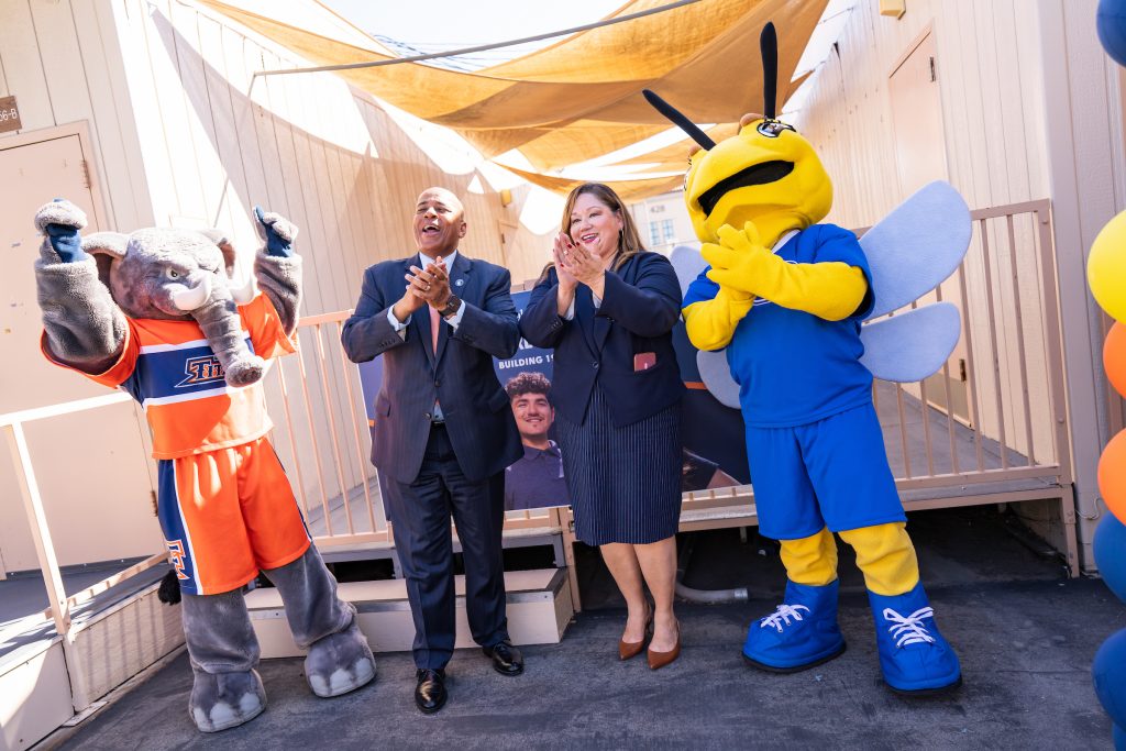 Two individuals in formal attire clap alongside two costumed mascots under a canopy at an outdoor event.