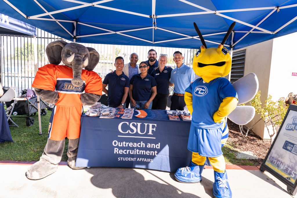 Two mascots and several staff members stand behind a CSUF Outreach and Recruitment table under a blue canopy.