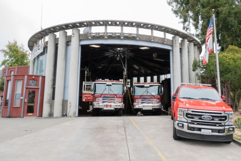 An exterior view of a round fire station's open garage, with two engines parked inside and a bright red pickup truck outside