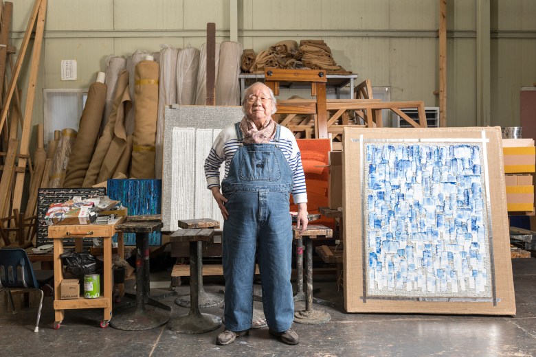 An older person in overalls stands in an art studio, surrounded by canvases, tools, and rolled fabrics.