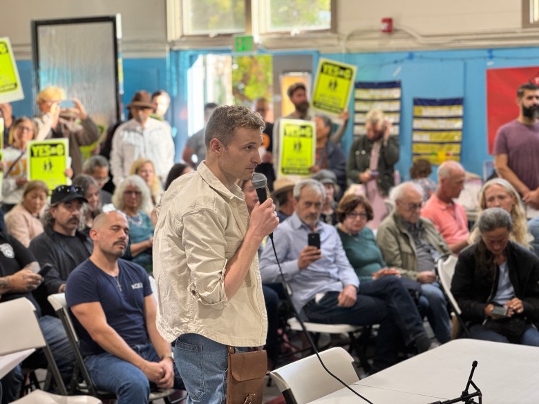 A person speaks into a microphone at a community meeting while people sit and stand, some holding yellow protest signs that read “YES.”.