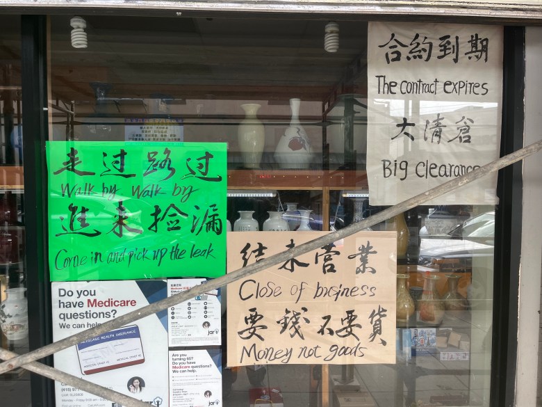 Storefront window with signs in English and Chinese announcing contract expiration, big clearance, close of business, and emphasizing “money not goods”; shelves with vases visible inside.