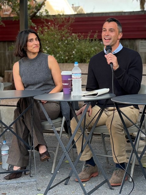 Two people sit at a table outdoors; the man on the right is speaking into a microphone while the woman on the left listens. Drinks and notebooks are on the table.