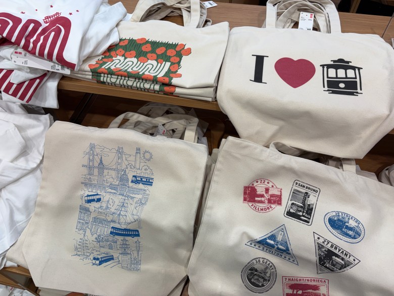 Four canvas tote bags on a store shelf, featuring designs of poppies, a cable car, a San Francisco city map, and various Muni transit-themed stamps.