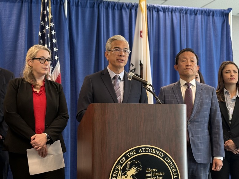 A man speaks at a podium labeled “Office of the Attorney General,” flanked by four people, with U.S. and California flags and a blue curtain in the background.