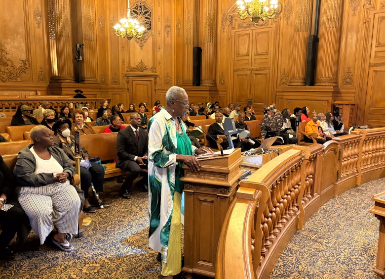 An older woman stands at a podium speaking in a formal wood-paneled room with seated audience members watching attentively.