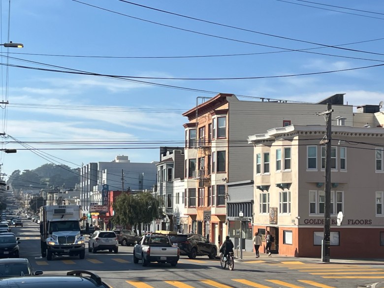 Street view of a busy intersection with cars, a cyclist, pedestrians, power lines, and multi-story buildings under a clear sky.