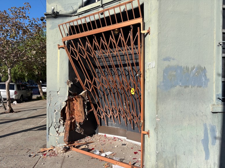 Damaged building exterior with a bent security gate and a hole in the wall near a doorway marked 2700.