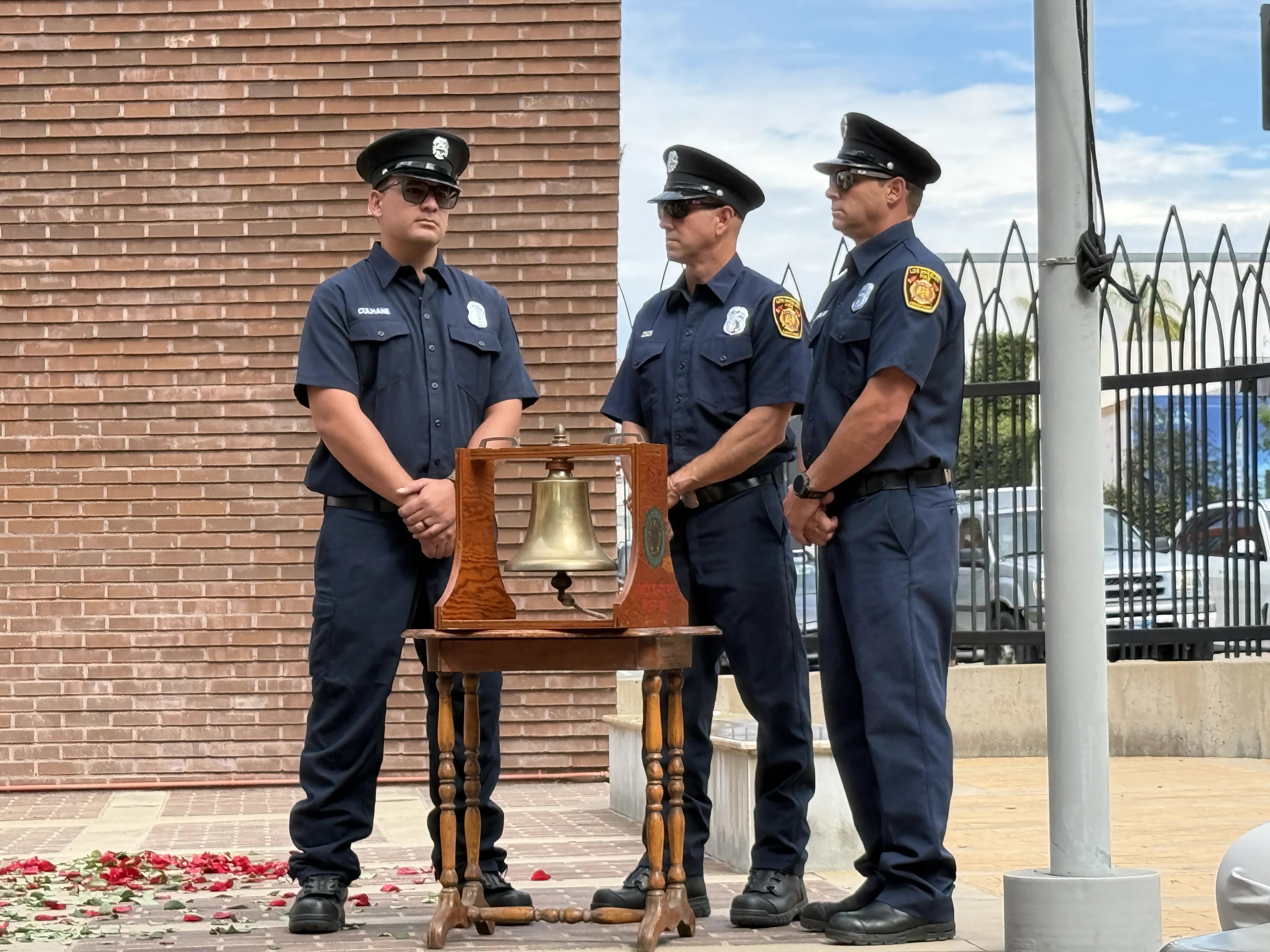 Three LAFD firefighter stand with their arms folded in from of them surrounding a brass bell mounted in a wooden case