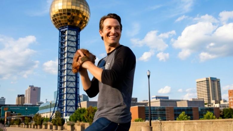 A smiling man in an AI-generated video, wearing workout clothes holds a football beside Knoxville’s Sunsphere, with downtown buildings under a bright blue sky; the scene feels upbeat and energetic.