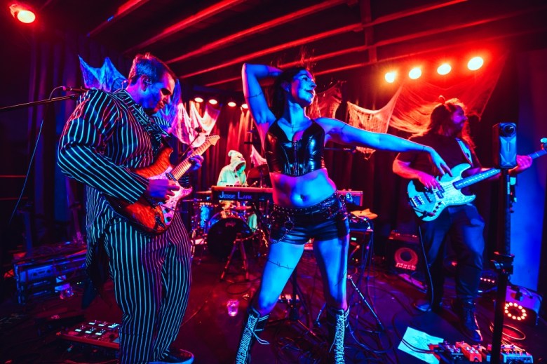 A band performs in costume, with a man playing guitar to the left and a woman posing at centerstage. A drummer can be seen wearing a hooded cape.