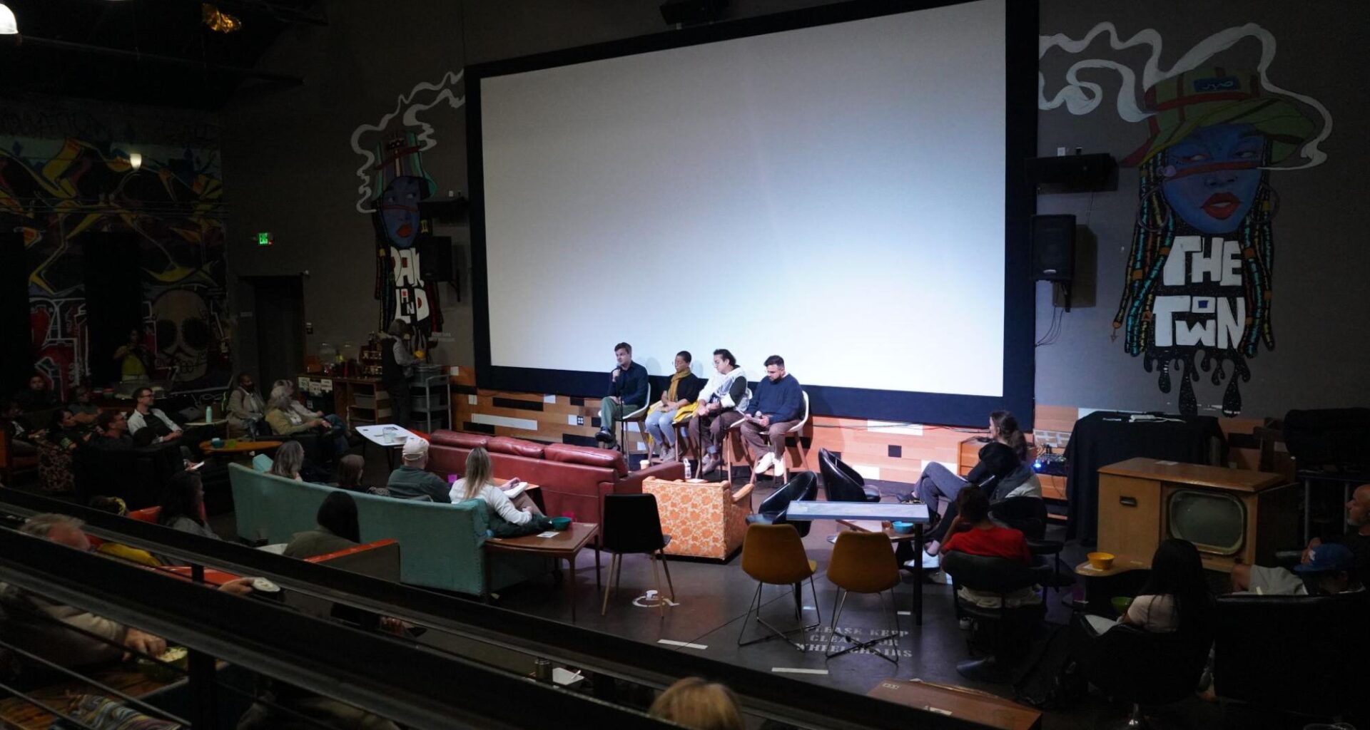 Four panel members sit in chairs on a stage in front of a large, blank projection screen while speaking into microphones. An audience sits in sofas and chairs facing the stage in a room decorated with wall murals, including text reading "The Town".