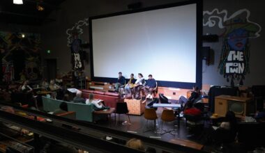 Four panel members sit in chairs on a stage in front of a large, blank projection screen while speaking into microphones. An audience sits in sofas and chairs facing the stage in a room decorated with wall murals, including text reading "The Town".