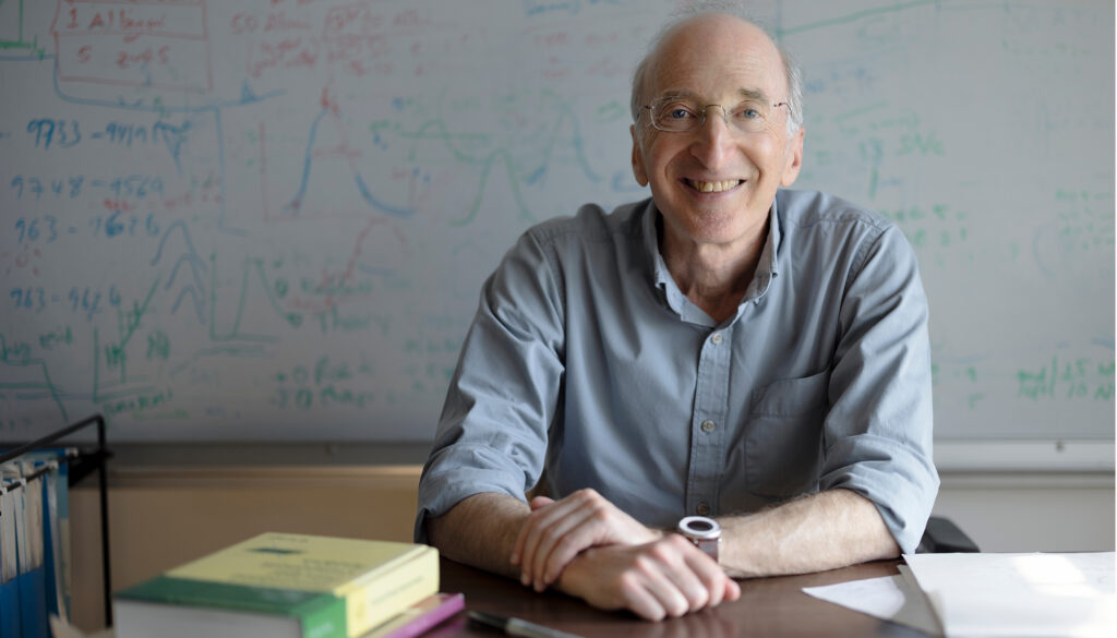 man in blue shirt and glasses at a desk in front of a whiteboard with colorful equations