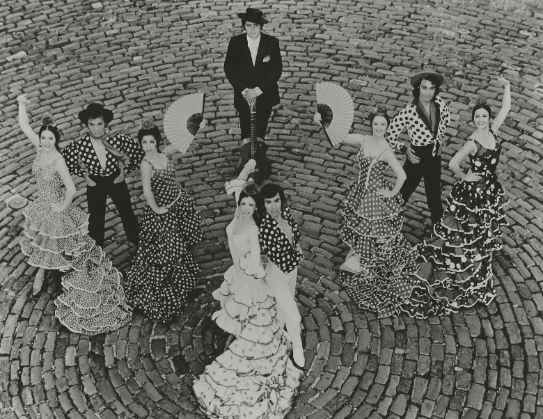 A group of seven flamenco dancers and a guitarist positioned in a circle on a cobblestone surface.