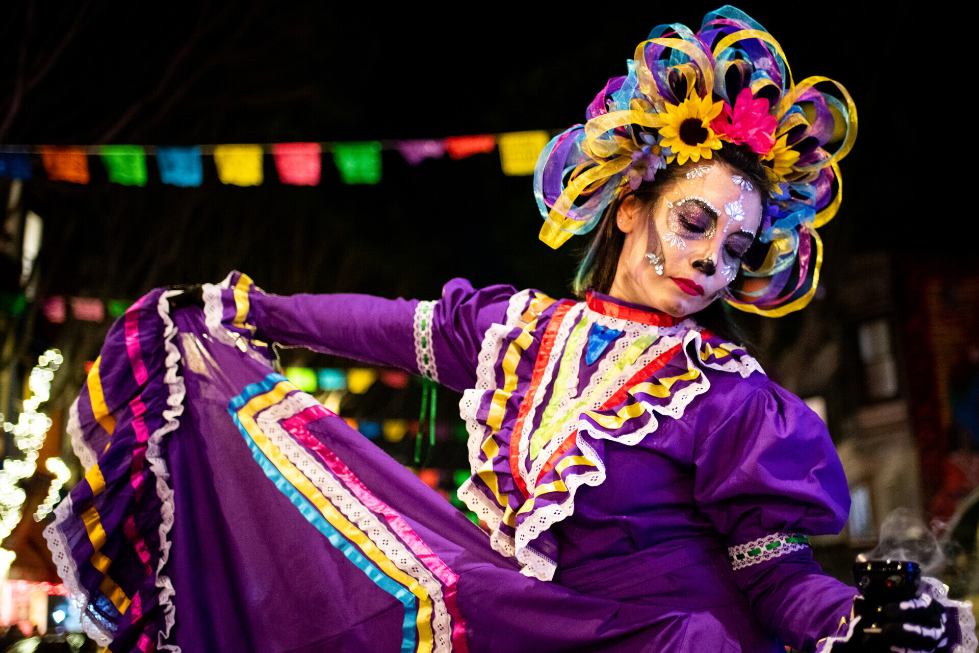 A person wears an elaborate headdress, face paint and purple, ribboned dress as they dance in the streets.