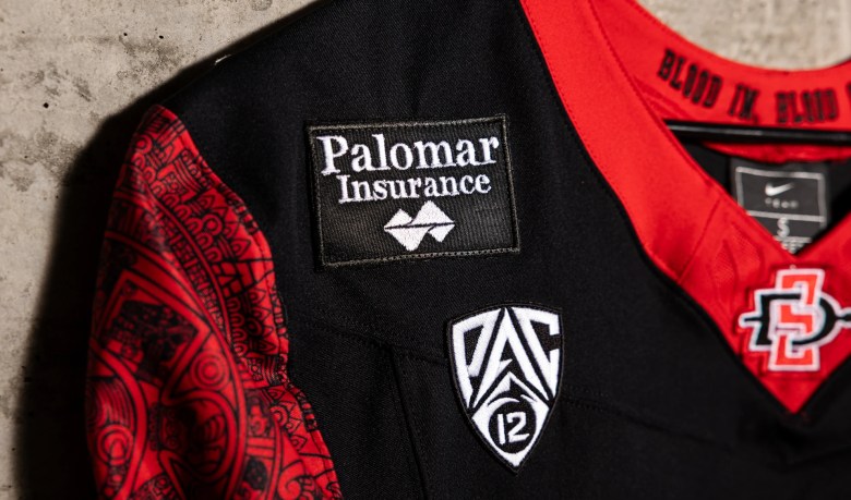 Close-up of a black-and-red athletic jersey with a Palomar Insurance patch, a white shield marked 12, and a red-and-white team emblem.