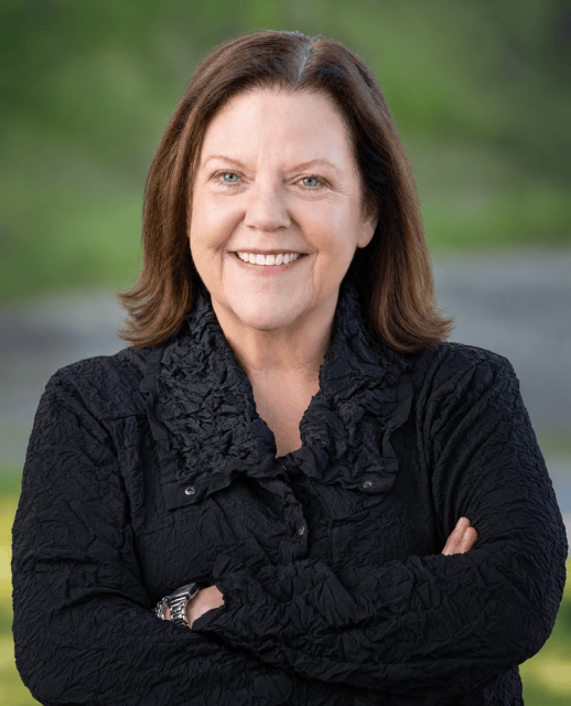 A smiling woman with shoulder-length brown hair, wearing a black collared top with her arms crossed.