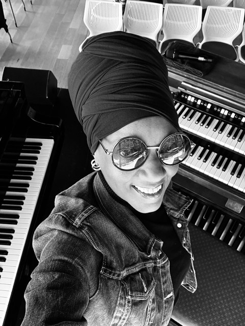 A smiling woman wearing a headwrap and round sunglasses takes a selfie between a piano and an organ.