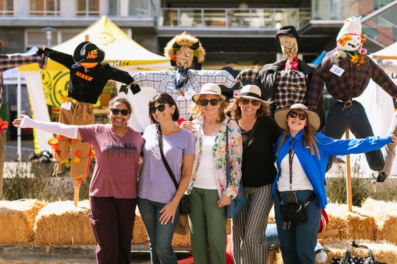 Five smiling women pose with arms outstretched in front of colorful scarecrows and hay bales at a sunny outdoor festival, creating a cheerful, playful scene.