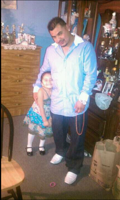 A man in a light blue striped shirt and a young girl in a patterned dress stand together in a room.