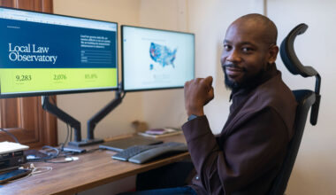 Diag Davenport, seated at a desk, with two computers screens showing his project. The screen on the left shows text that says