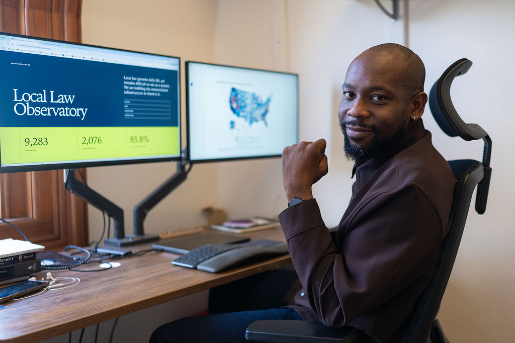 Diag Davenport, seated at a desk, with two computers screens showing his project. The screen on the left shows text that says