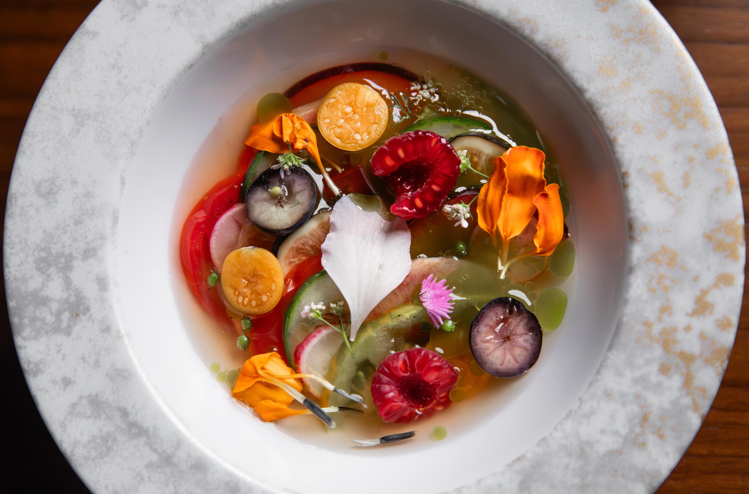 A bowl of cut fruit, such as raspberries, grapes, and tomatoes, alongside slices of cucumber and radish, in a clear broth at Shinka in San Francisco.