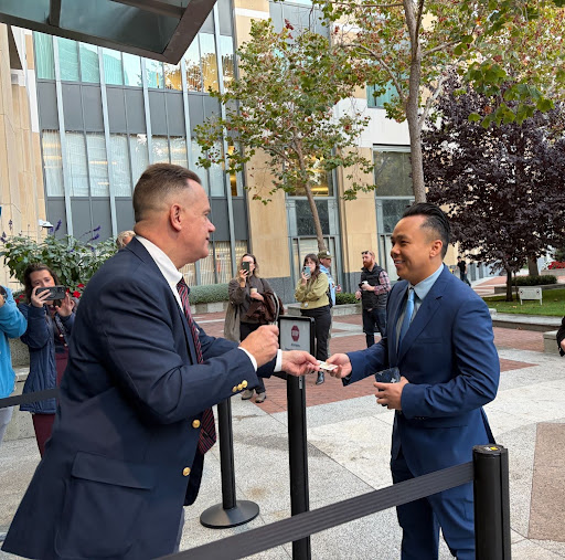 Outside a tall glass and stone paneled building, about half a dozen people aiming cameras at two men in navy blue suits. One man is handing an ID card to the other man at the entrance of the building.